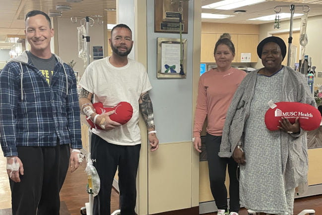Four people stand in a hospital waiting room, some with IVs attached. They are holding plush pillows shaped like kidneys with the 168[content00inject] Health logo printed on them.